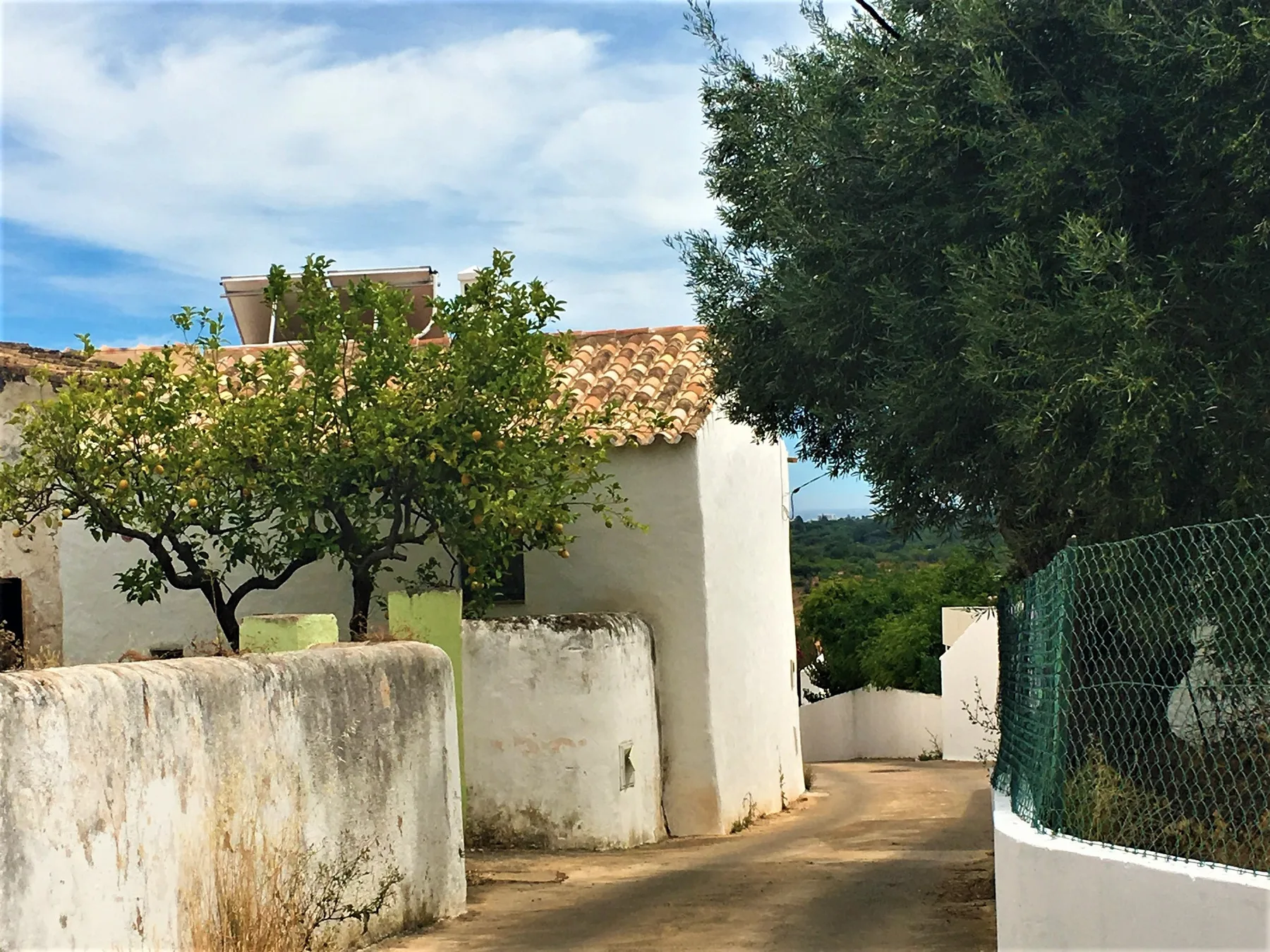 Una casa blanca se encuentra junto a un camino estrecho, con árboles y un tejado de tejas.