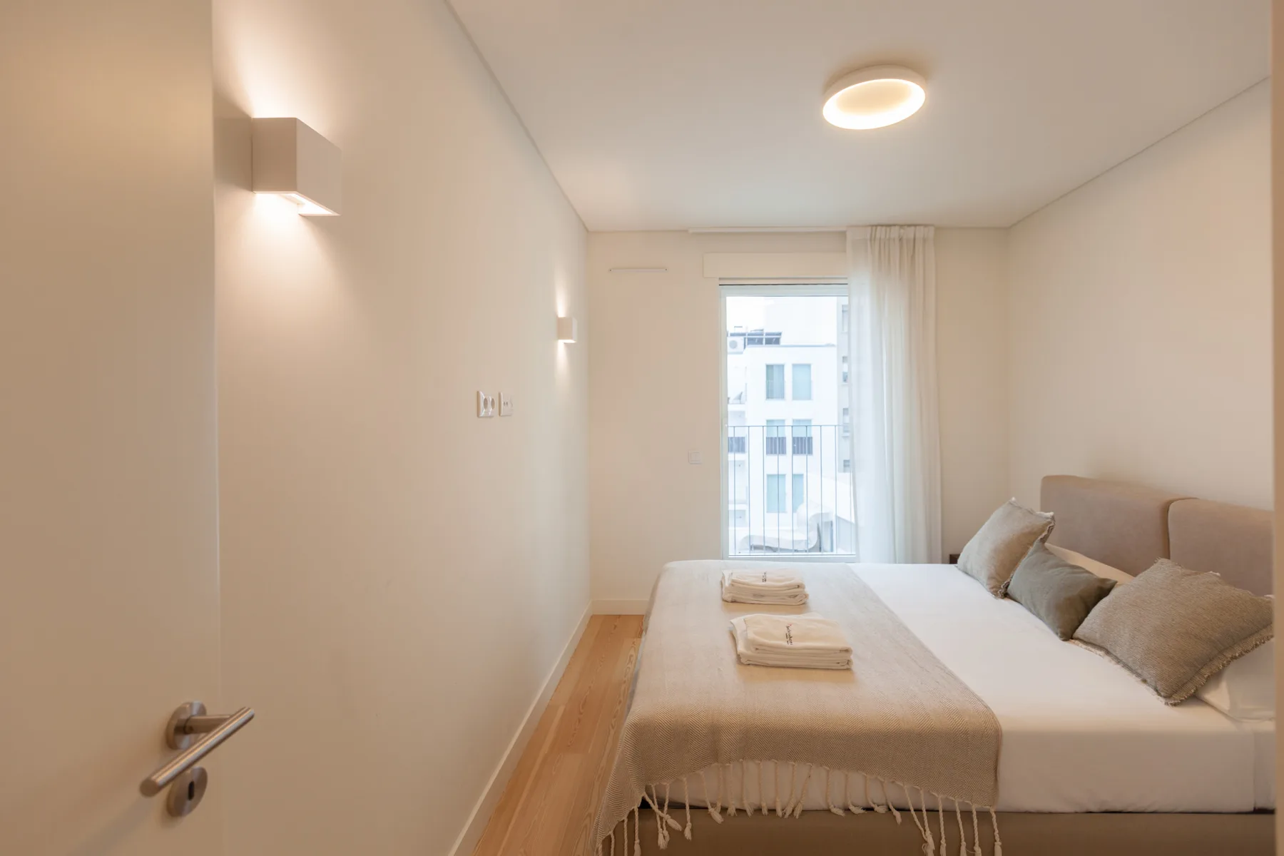 Un dormitorio con una cama, iluminación junto a la pared y una puerta de cristal que da a un balcón.