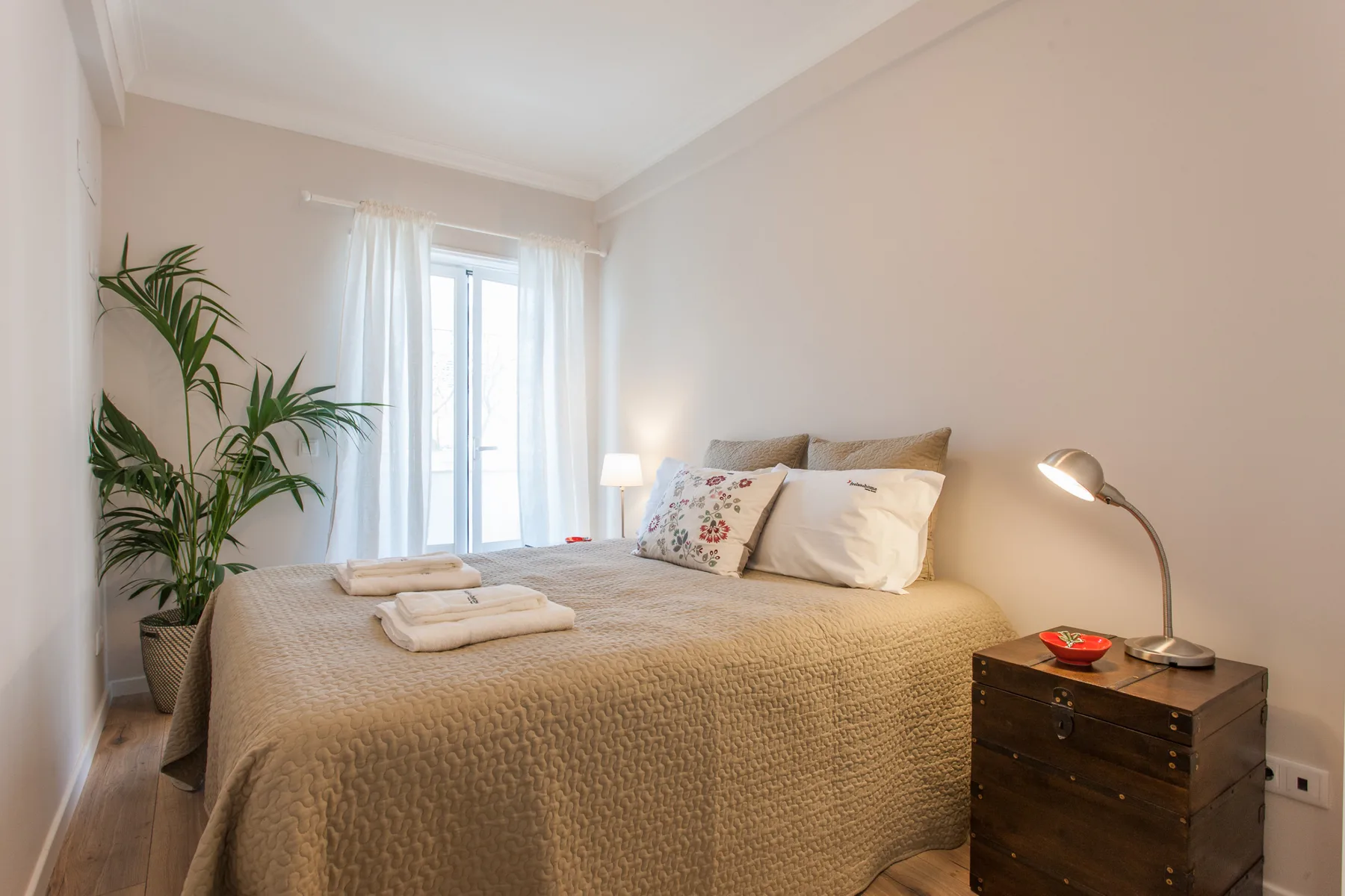 A bright bedroom with a bed, bedside table, and a window with sheer curtains.