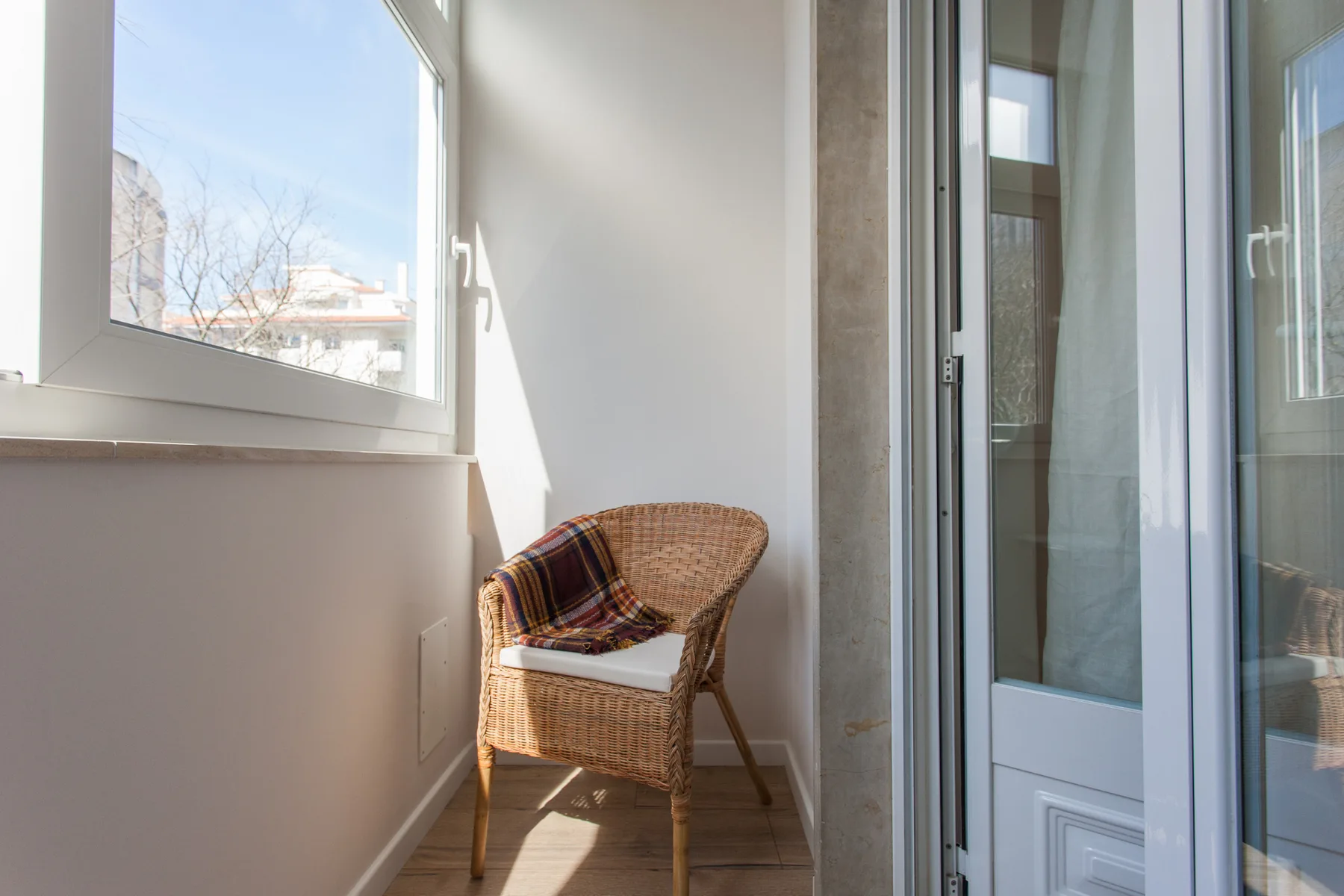 A bright enclosed balcony with a wicker chair beside large windows.
