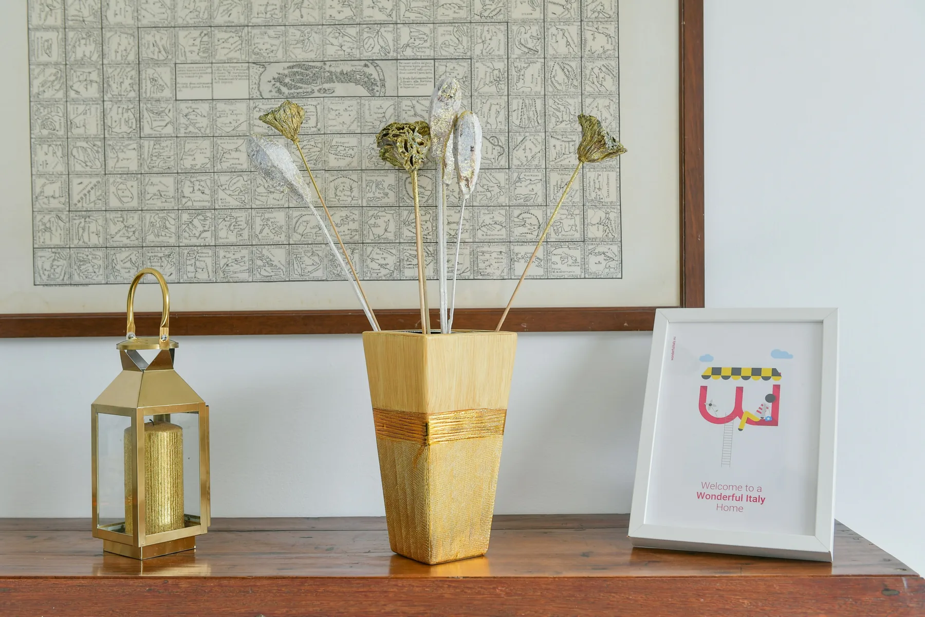 Una consola de madera exhibe un jarrón decorativo, un farol y una lámina enmarcada.