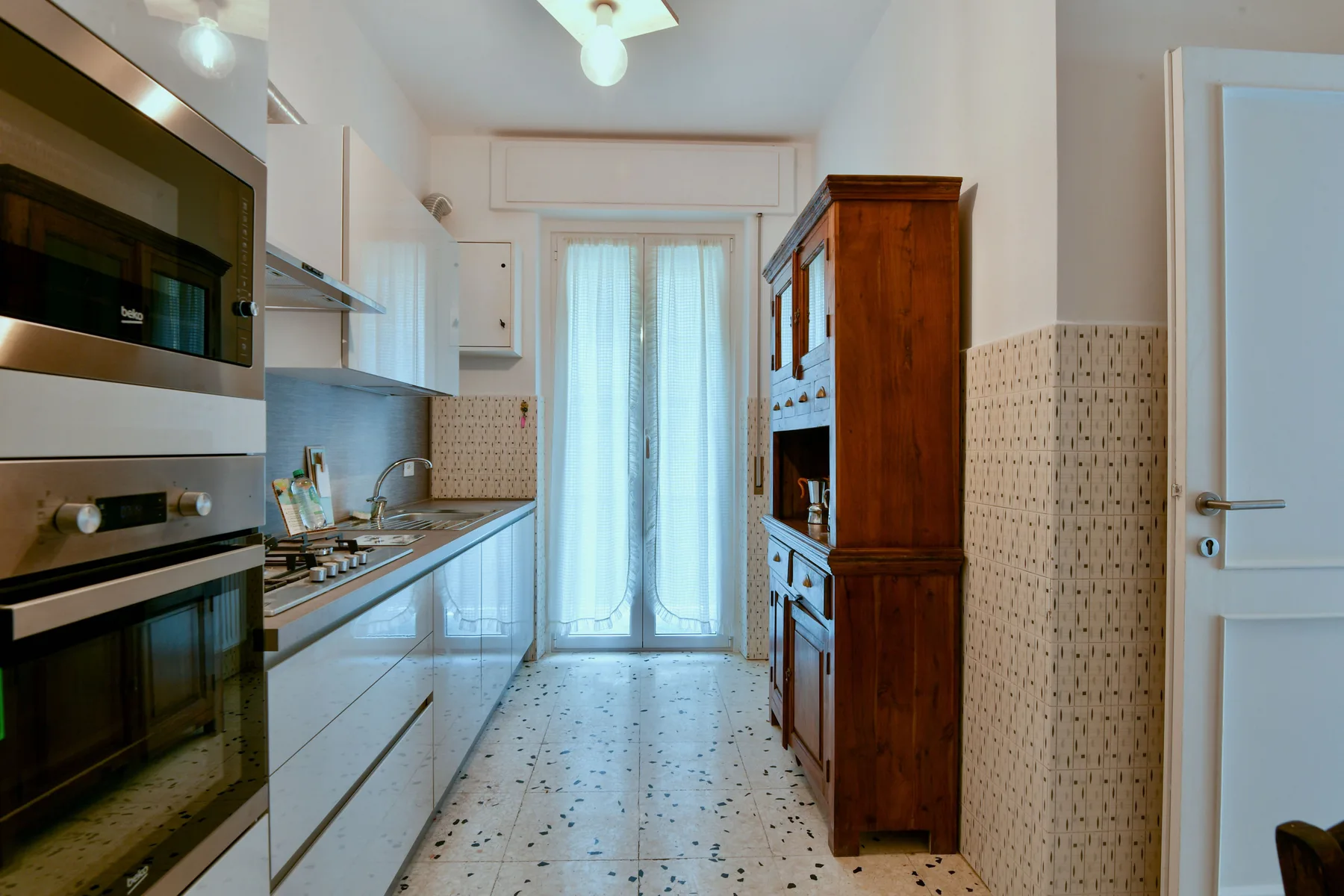 Una cocina estrecha con armarios blancos, una placa de cocina y un armario alto de madera.