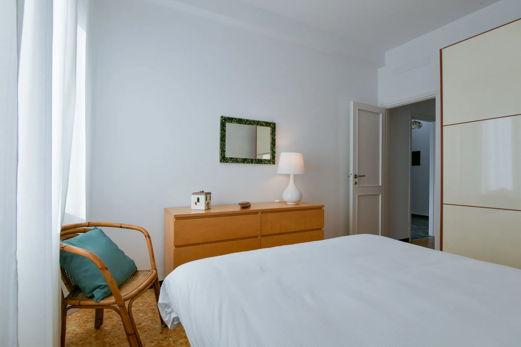 Un dormitorio con una cama tendida, una cómoda y una silla junto a una ventana luminosa.