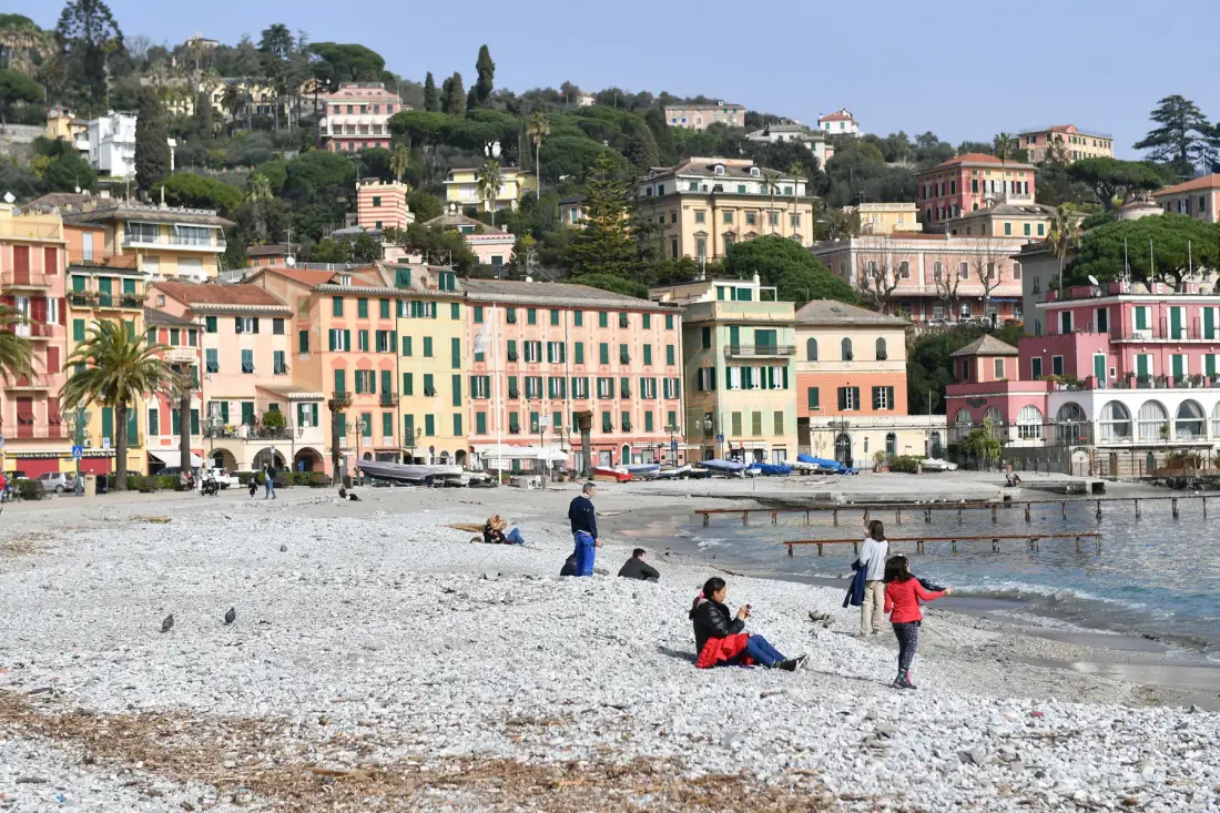 Una playa de guijarros junto al paseo marítimo, con edificios coloridos en la ladera.