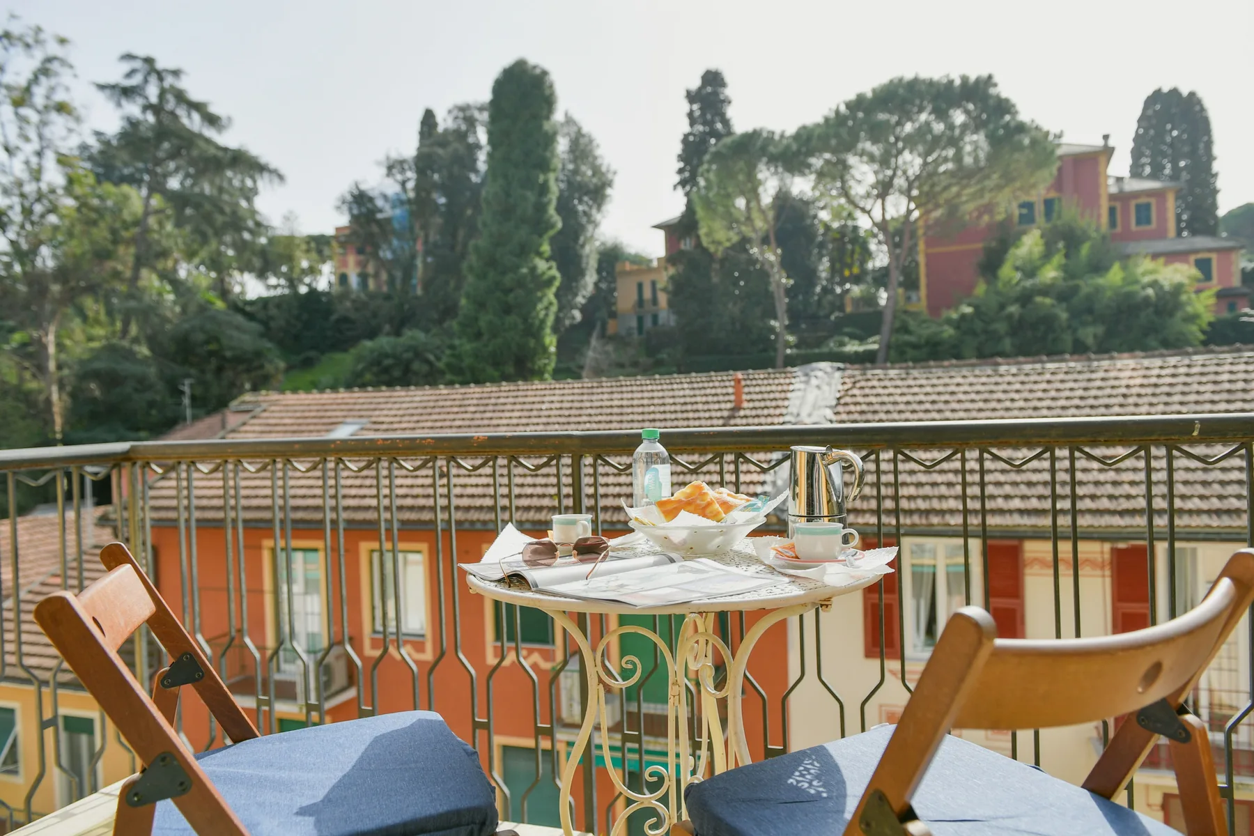 Una mesa de comedor en un balcón, con platos y bebidas, da a tejados y árboles cercanos.