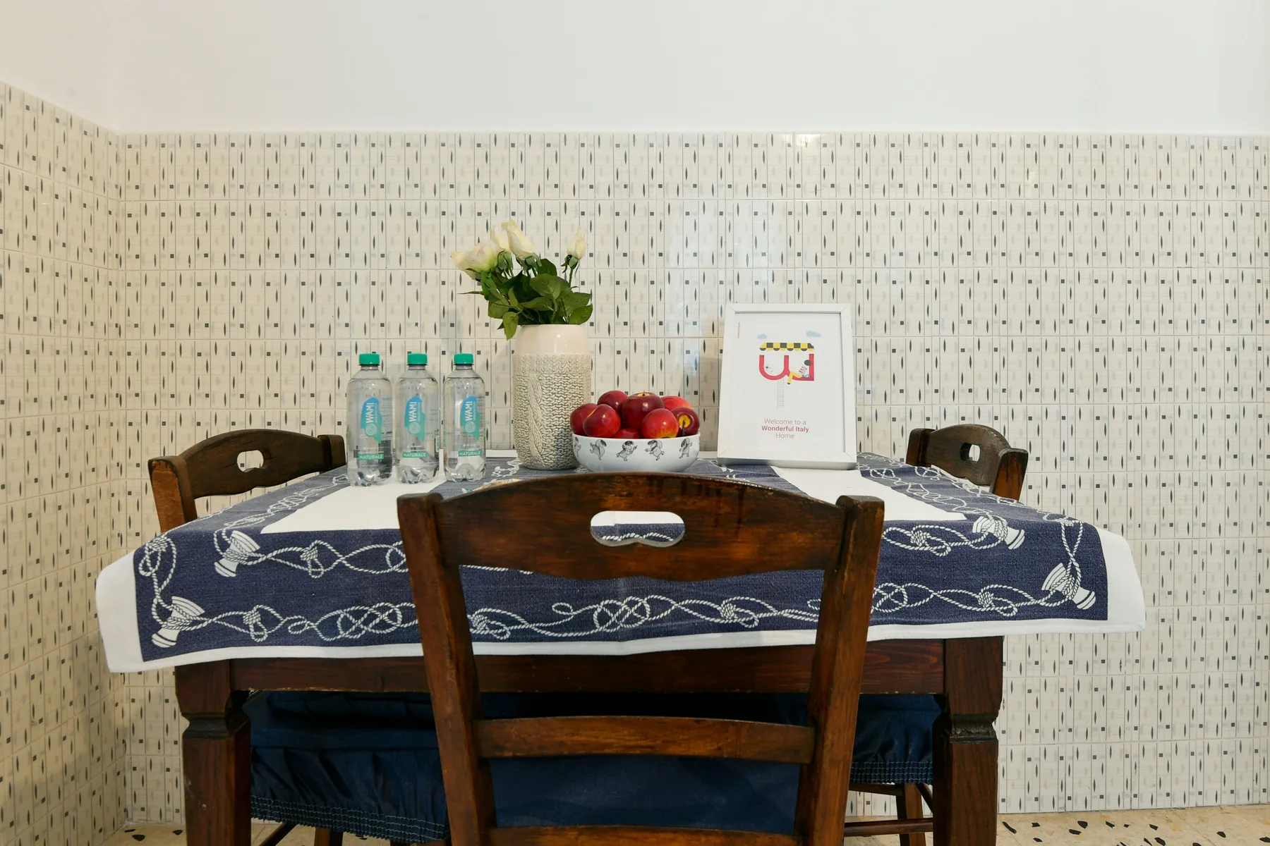 Una mesa de comedor con un camino estampado, fruta y flores está junto a una pared de azulejos.