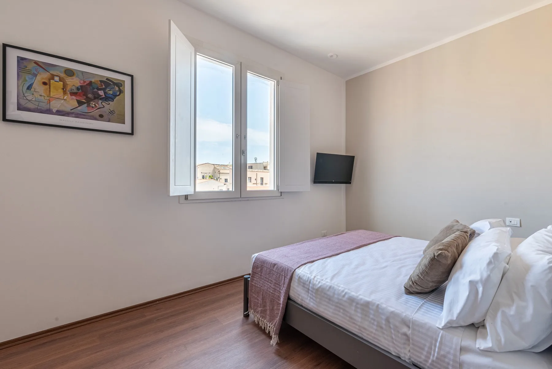 Un dormitorio luminoso con una cama, ventanas abiertas y un televisor en la pared.