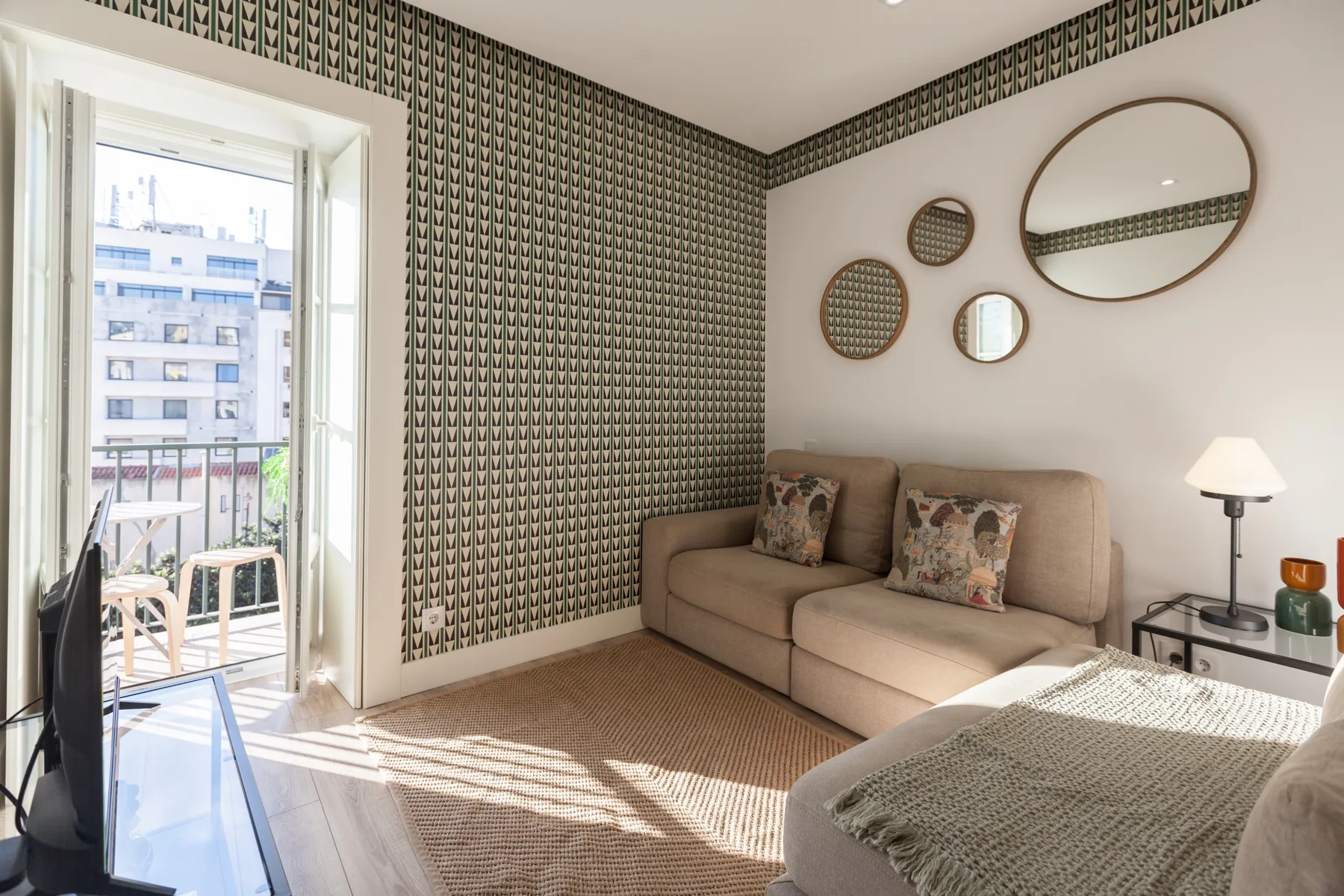 A living room with a beige sofa, patterned wallpaper, and round wall mirrors.