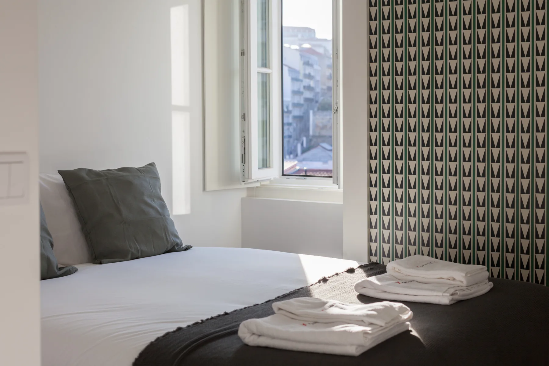 A bedroom with white bedding beside an open window and patterned curtains.