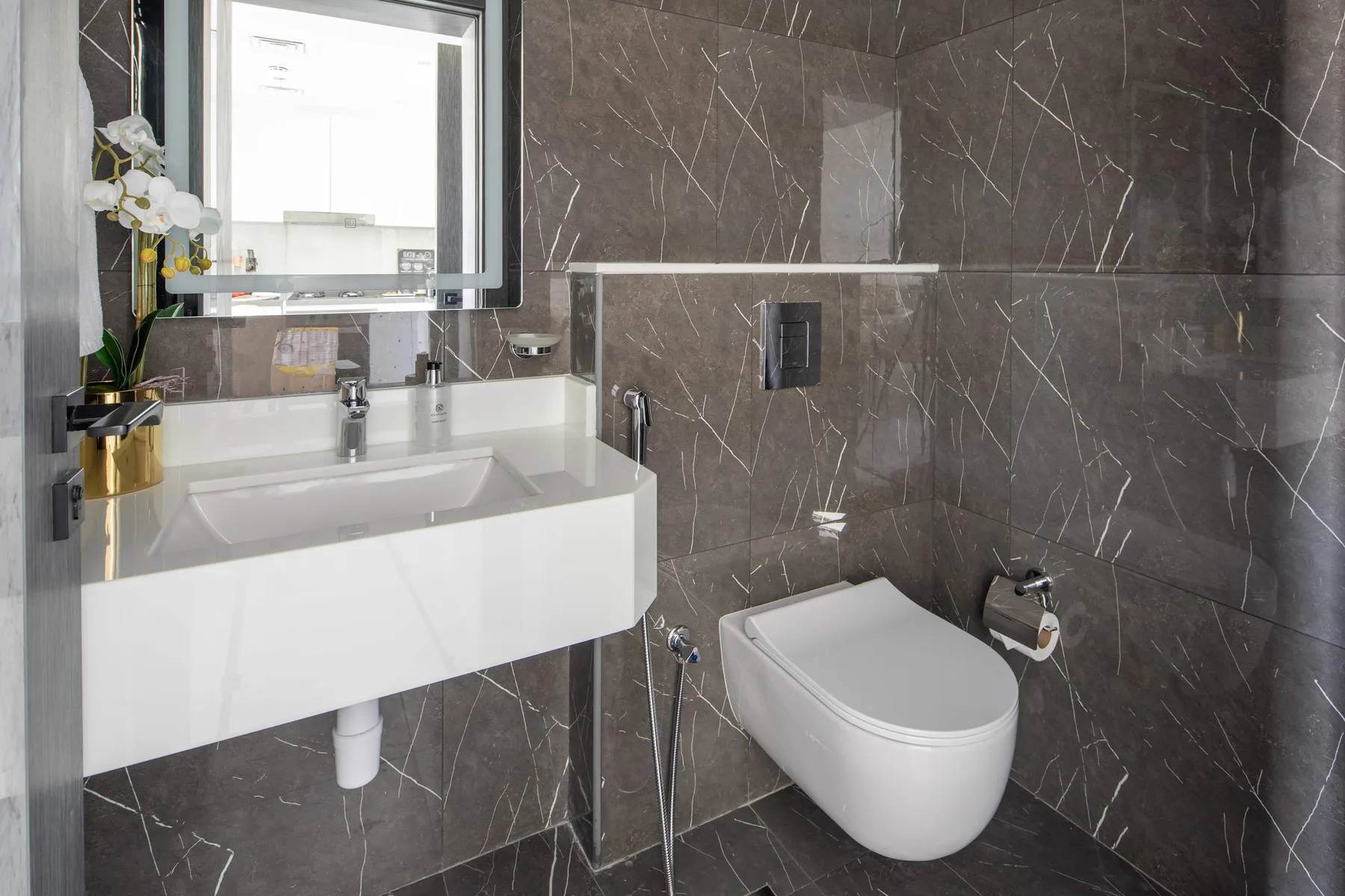 A modern bathroom with a wall-mounted sink and toilet against dark tiled walls.