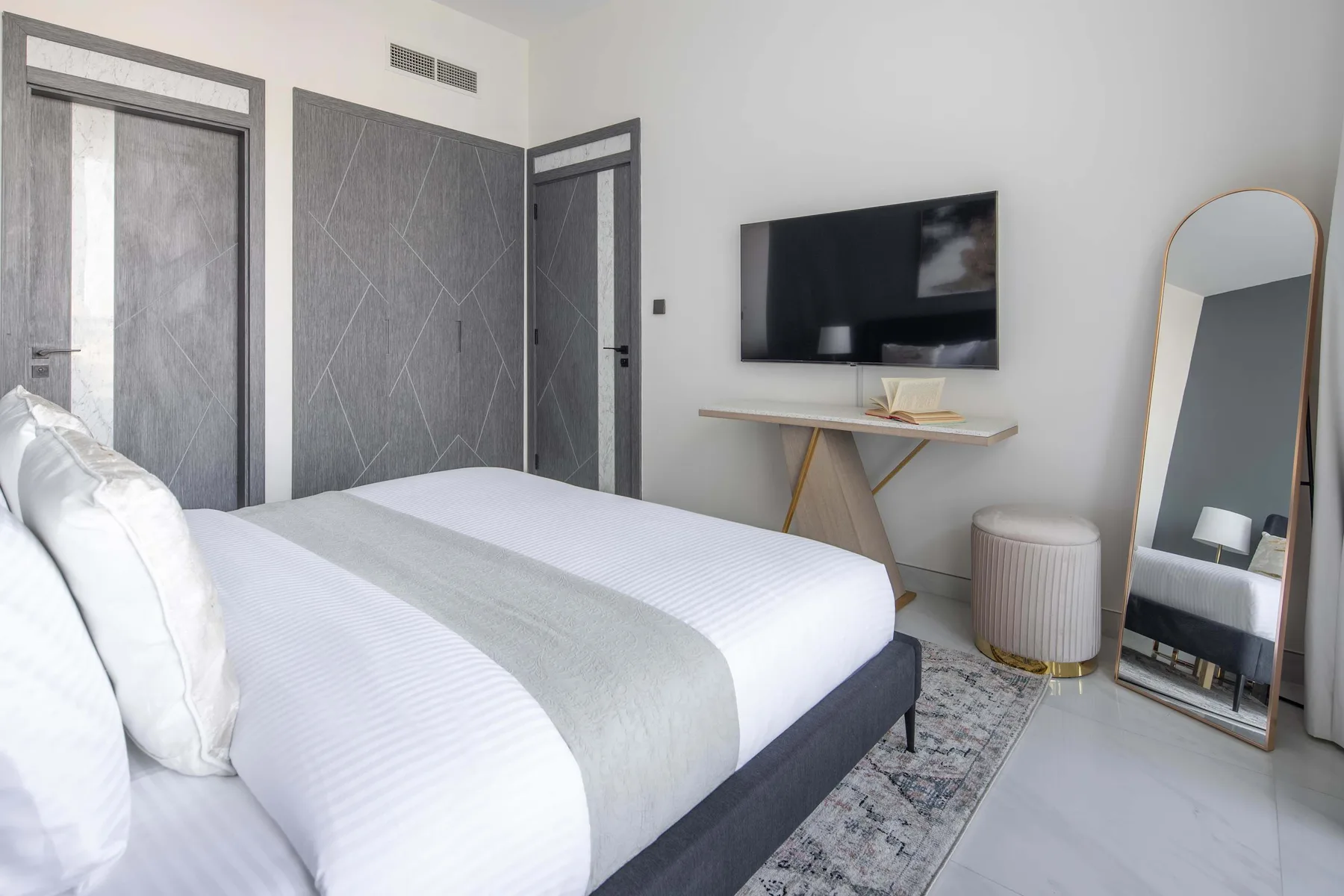 A bedroom with a neatly made bed, wall-mounted television, and full-length mirror.