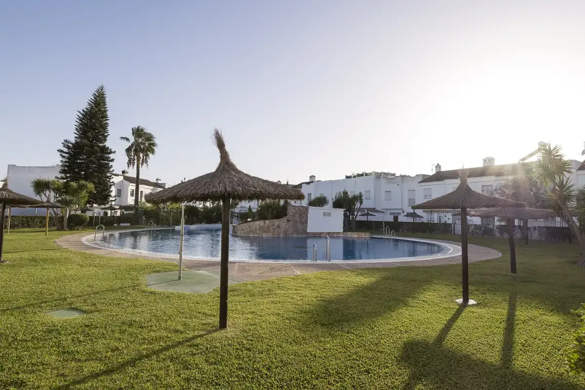 An outdoor swimming pool sits among grassy lawns and thatched shade canopies.