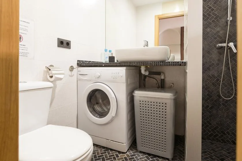 A bathroom with a washing machine, laundry hamper, toilet, and shower.