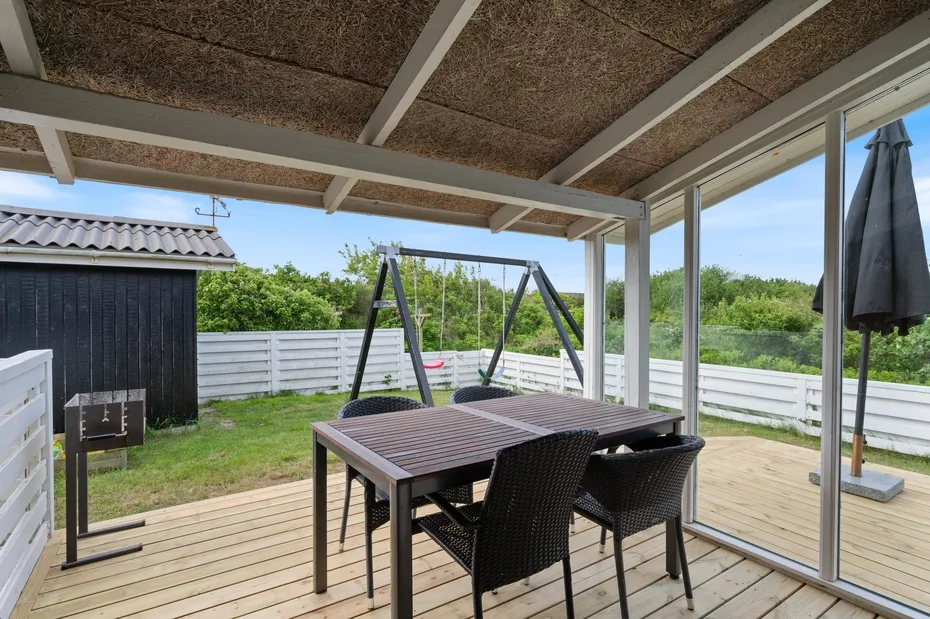 A covered outdoor deck with a dining table and seating overlooks a fenced yard.