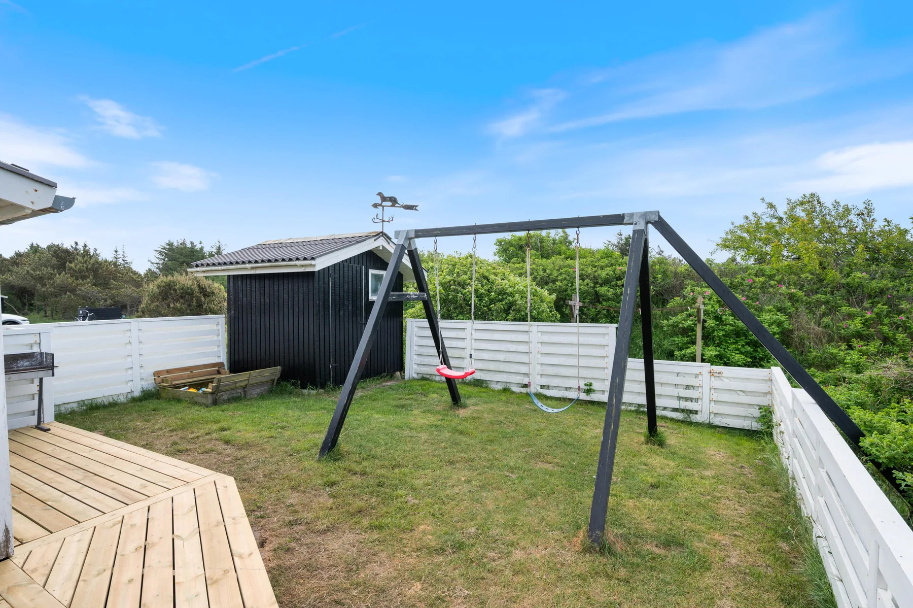 A fenced backyard with a wooden deck, shed, and swing set under a clear sky.