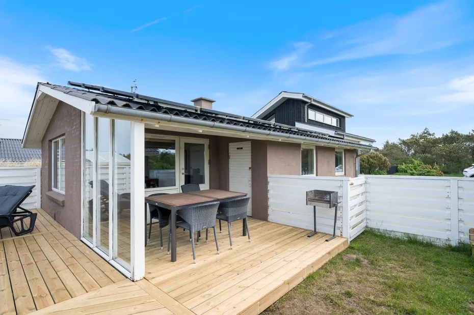 A house opens onto a wooden deck with outdoor dining furniture and a grill.