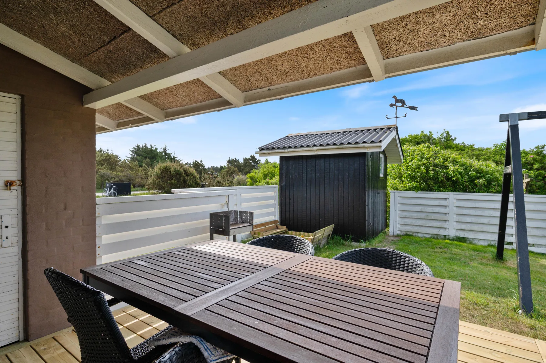 A covered outdoor dining area with a table, seating, and a fenced yard.