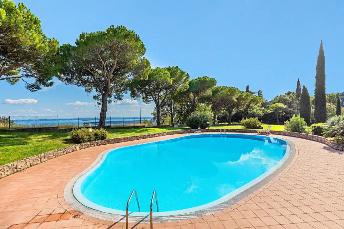 An outdoor swimming pool sits among trees, with the sea visible beyond the garden.