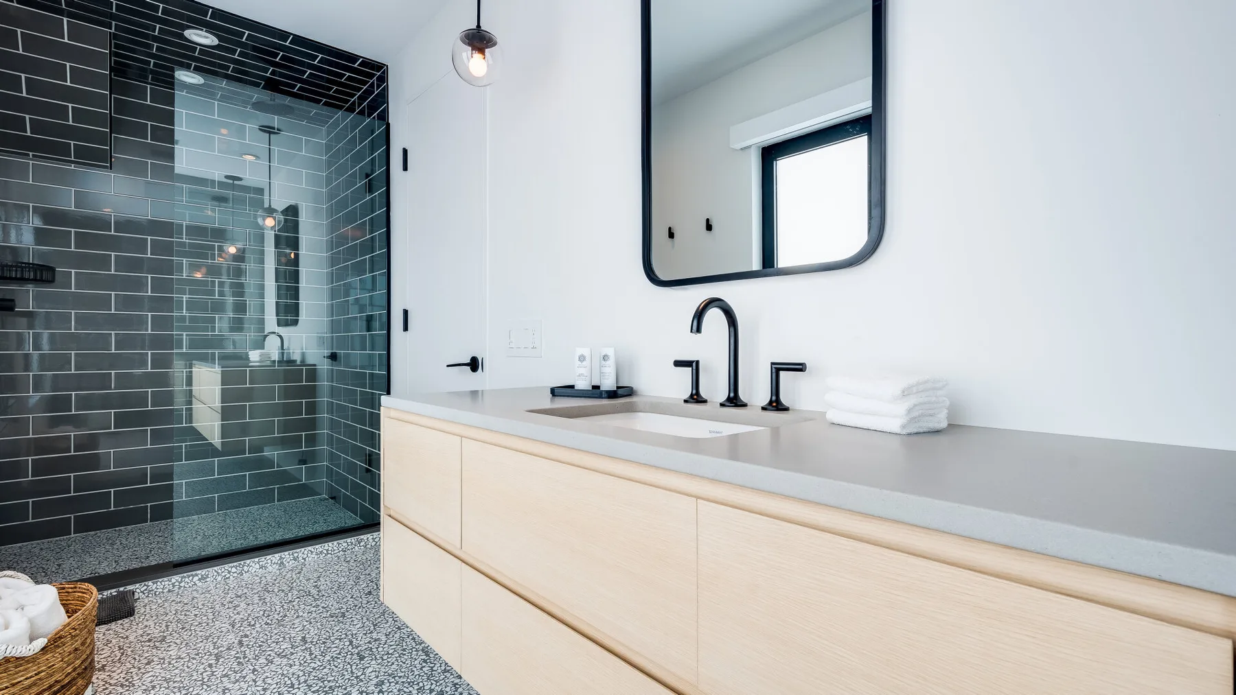 A bathroom with a glass shower, dark tiled walls, and a light wood vanity.