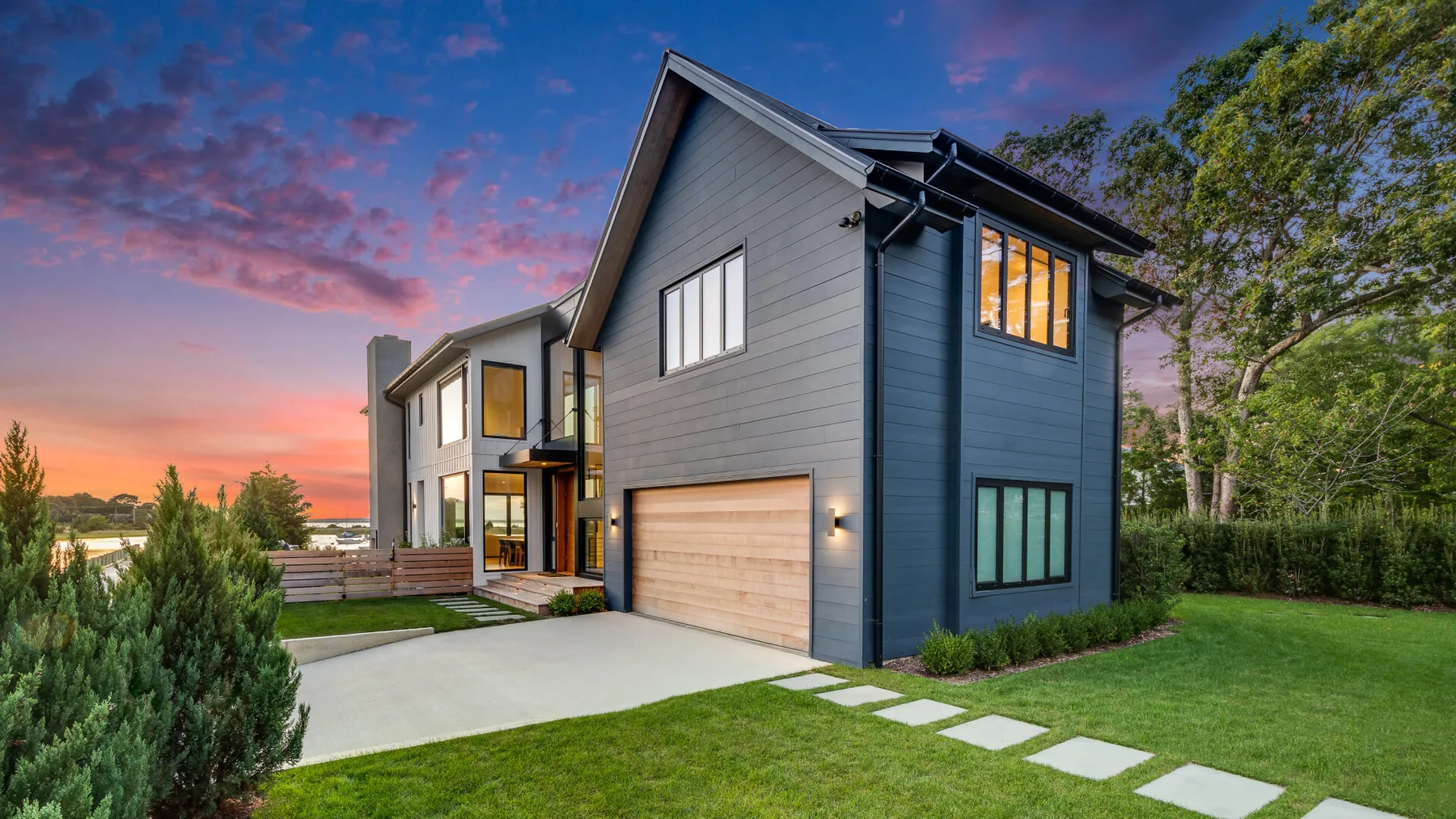 A modern house stands beside a driveway and green lawn at sunset.