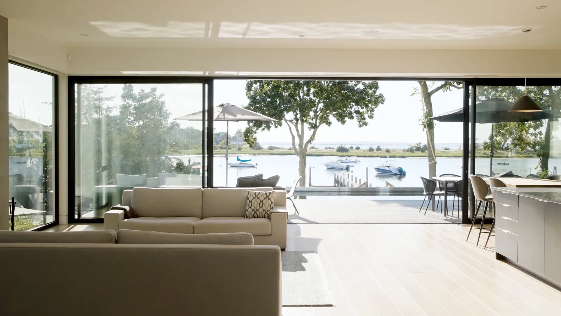 A bright living room opens onto a waterfront patio, with wide glass windows.