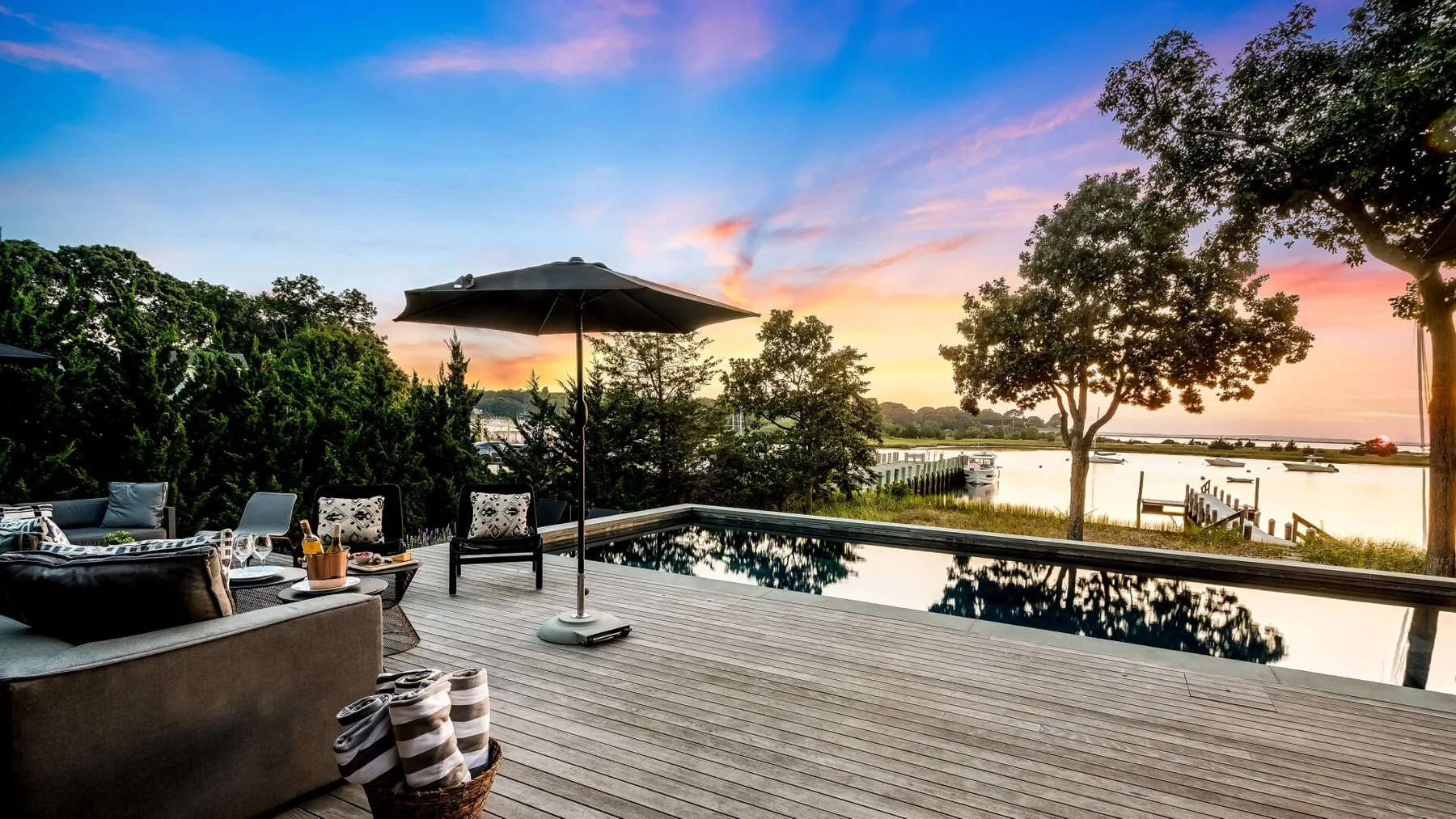 An outdoor poolside seating area overlooks a calm body of water at sunset.