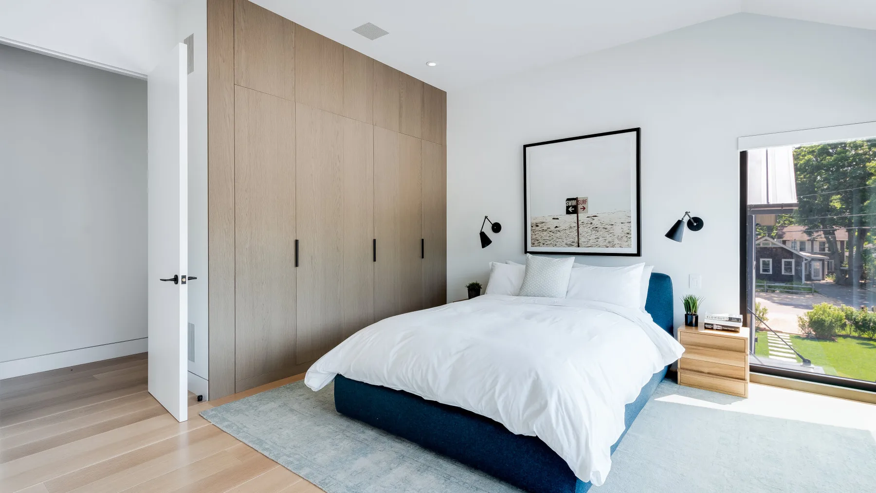 A bright bedroom with a white bed, wooden wardrobes, and a large window.