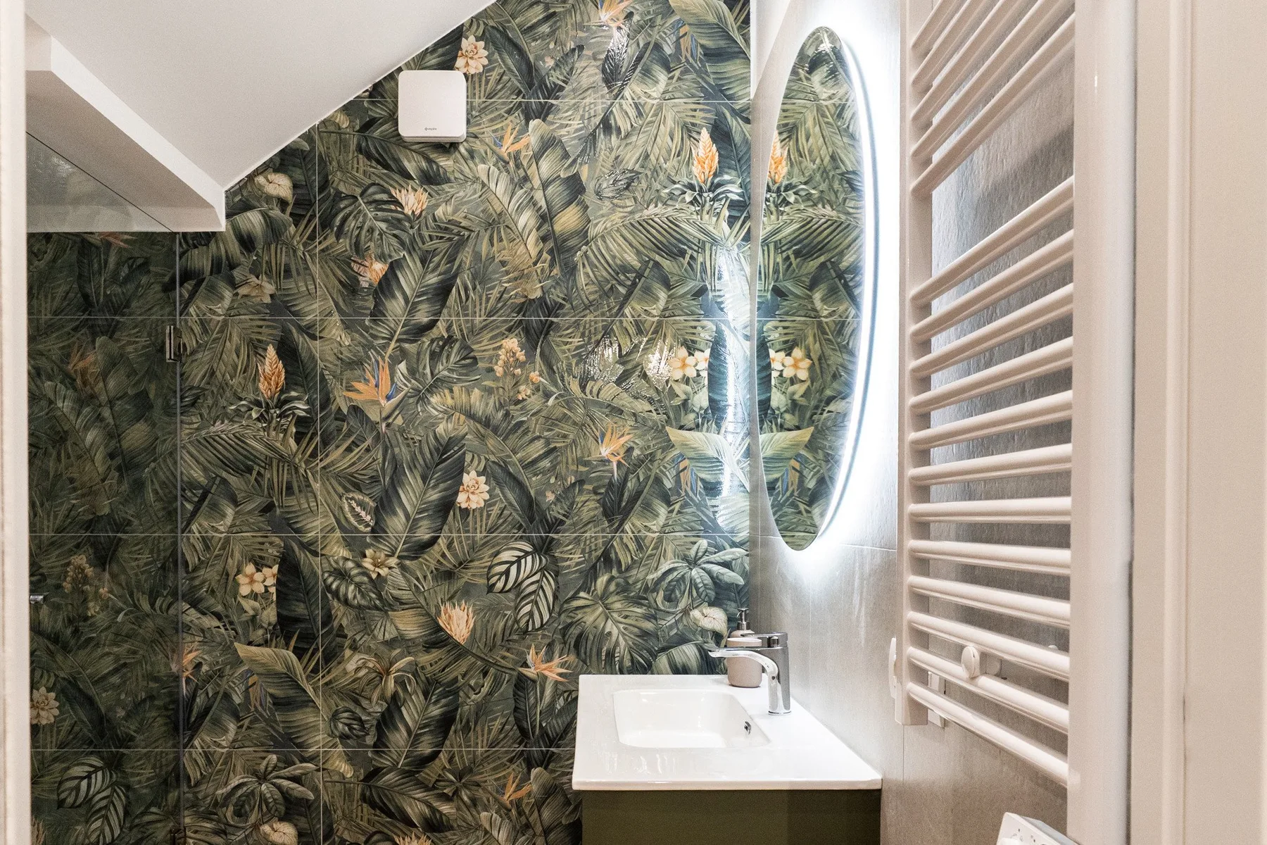 Un baño con lavabo, espejo iluminado y una pared con estampado tropical.
