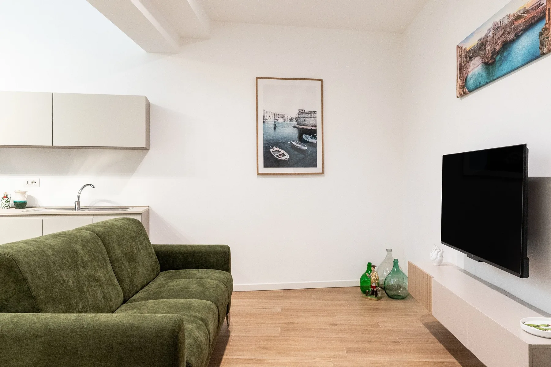 Una sala de estar con asientos verdes, un televisor de pared y una cocina compacta.