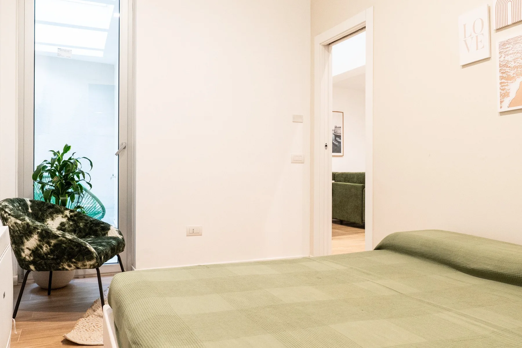 Dormitorio con una colcha verde y una silla estampada junto a la ventana.