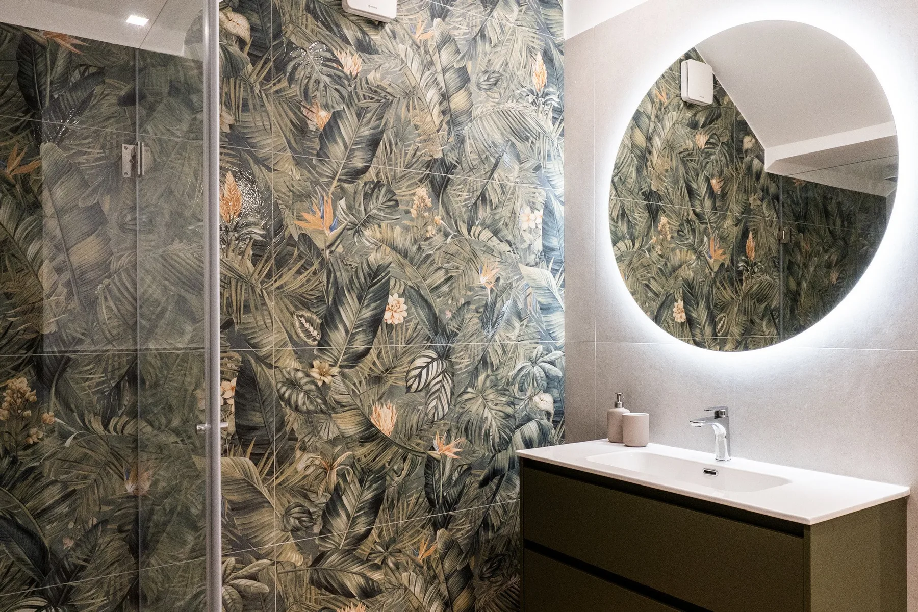 Un baño con ducha de vidrio, paredes con motivos de hojas y un espejo redondo sobre el lavabo.