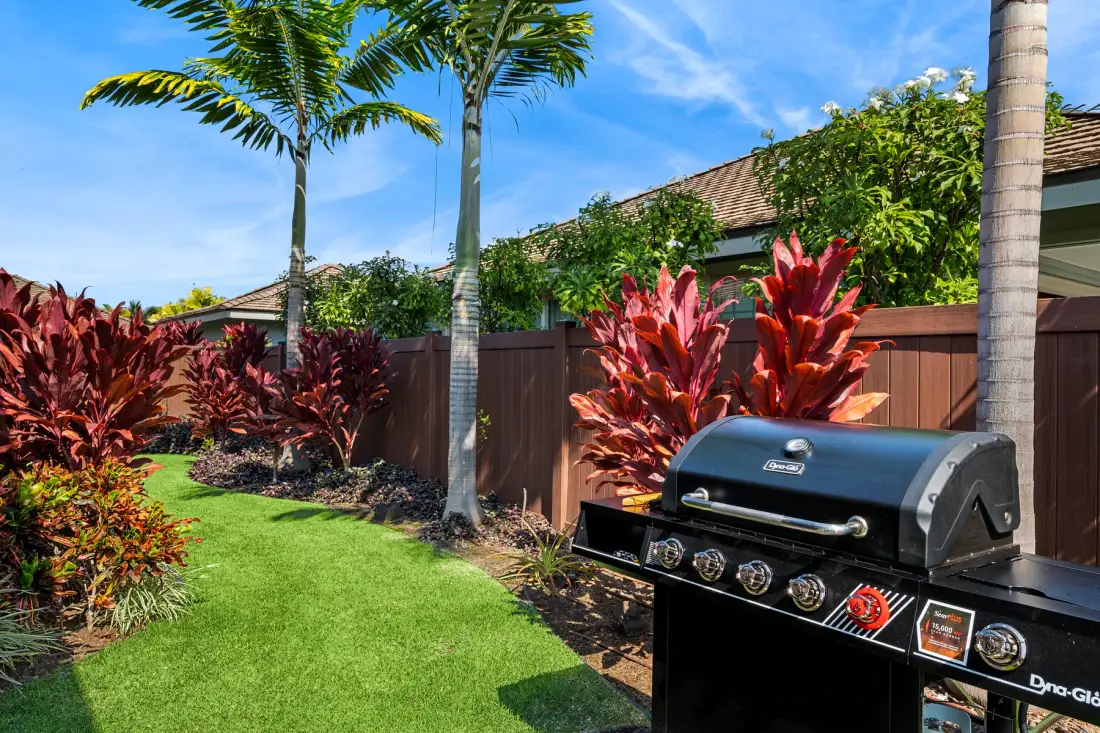 Un jardín con plantas tropicales y una parrilla junto a una cerca de madera.