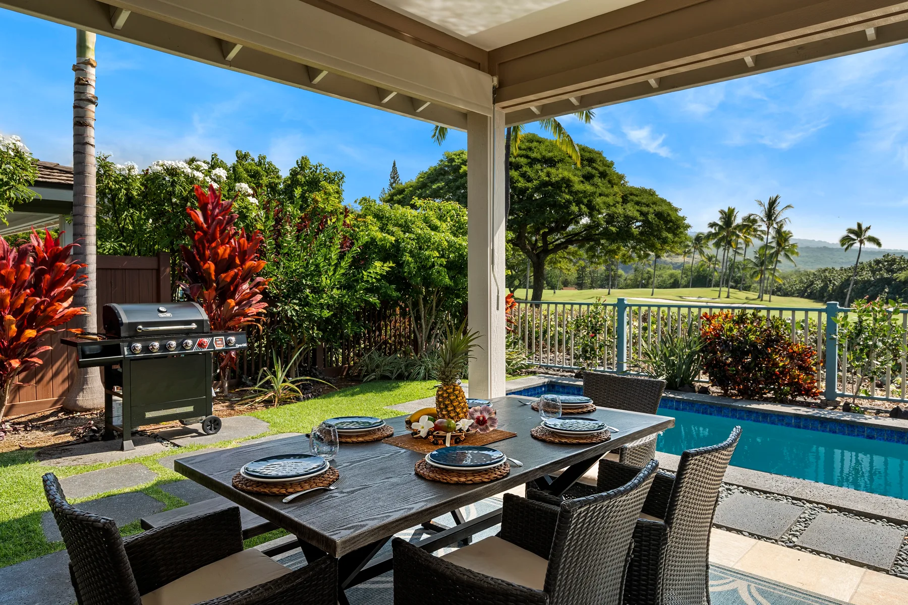 Una zona de comedor exterior cubierta da a una piscina, un jardín y colinas lejanas.