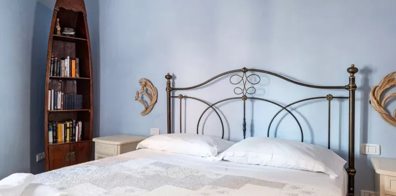 Un dormitorio con una cama de metal, ropa de cama blanca y una estantería alta.