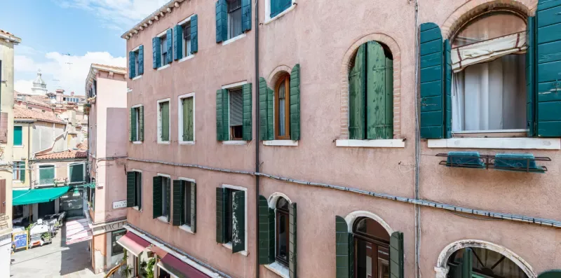 Un edificio rosa de varias plantas con contraventanas verdes junto a una calle estrecha.
