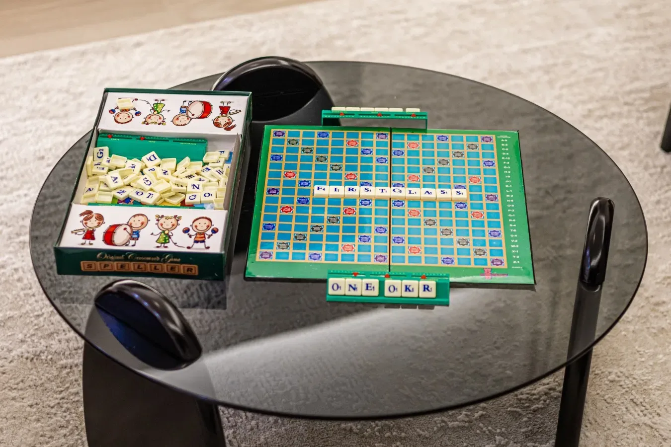 Un juego de mesa está dispuesto sobre una mesa redonda de vidrio junto a una caja de fichas con letras.