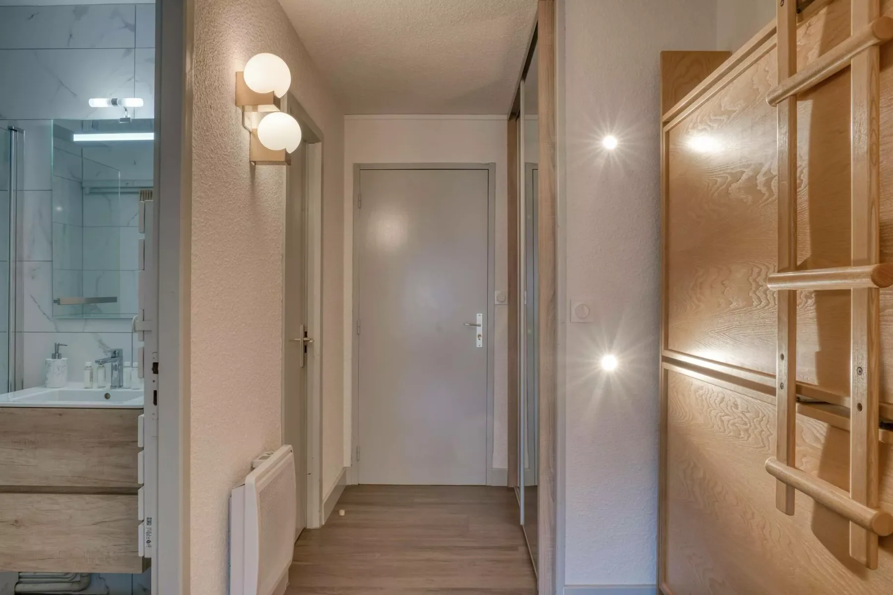 A hallway with a closed door, wall lights, and a bathroom visible to the side.