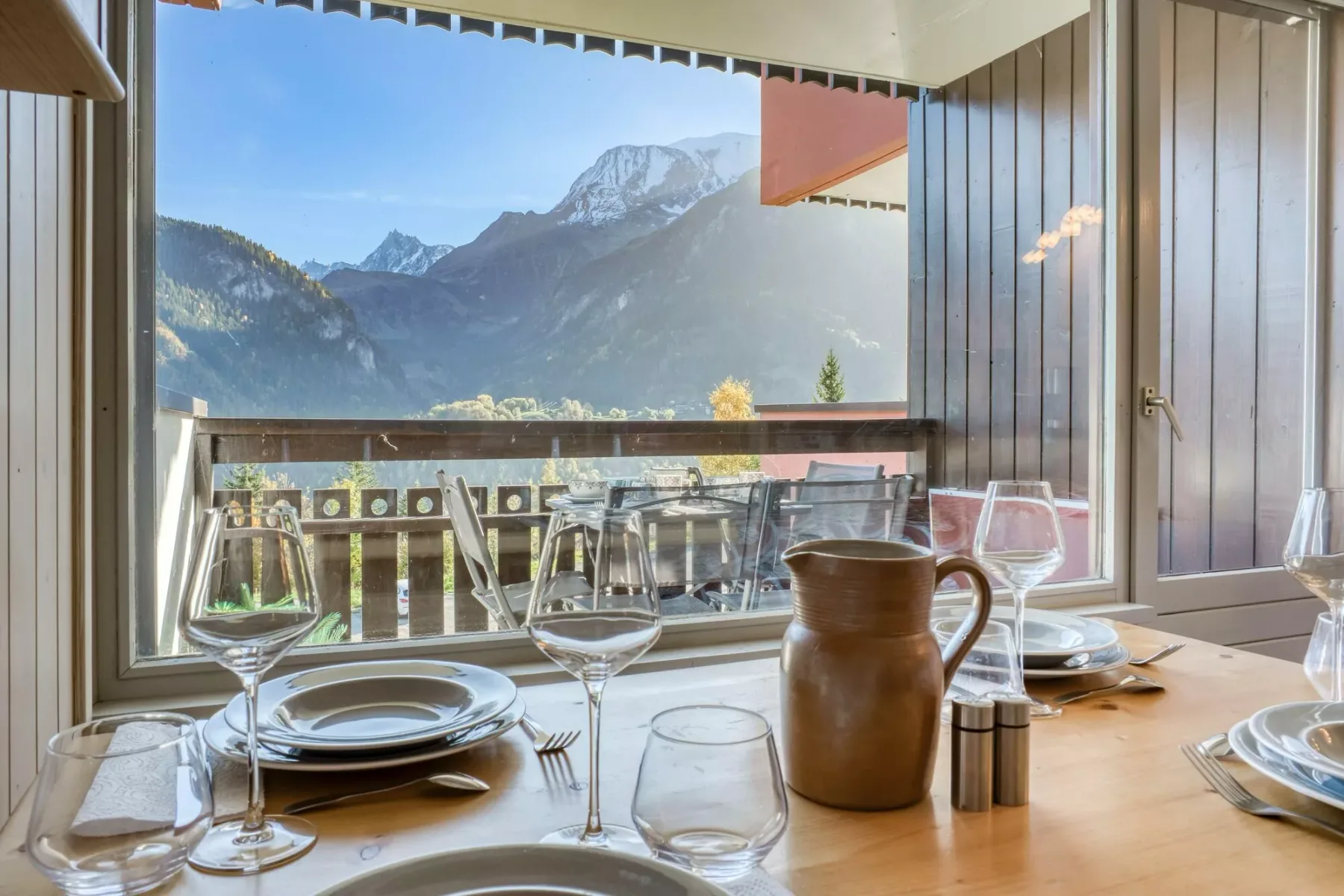 A dining table is set beside glass doors, with mountain views beyond the balcony.