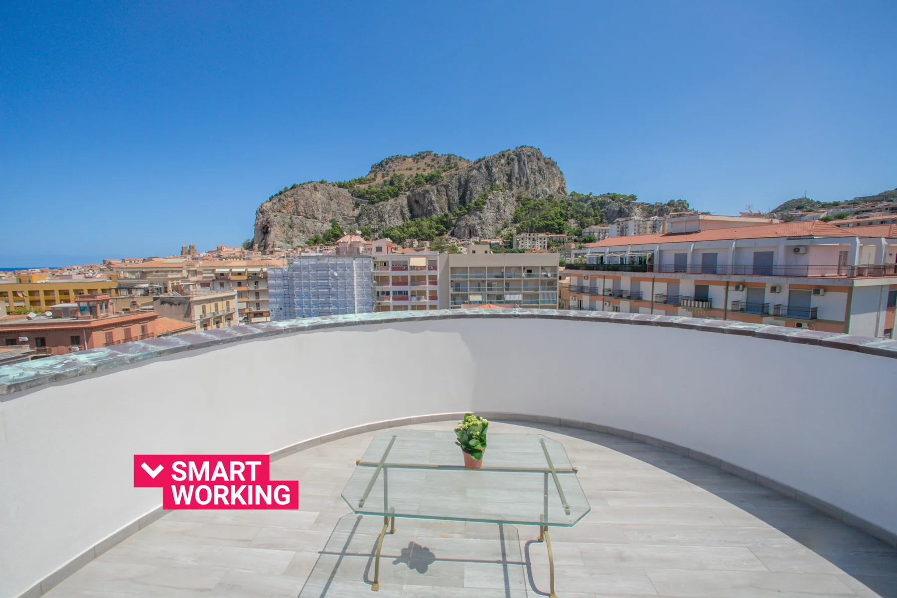 A rooftop terrace with a glass table overlooks nearby buildings and a rocky hill.