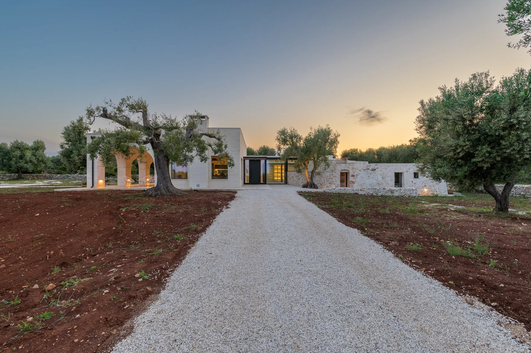 A modern white building stands at the end of a gravel driveway among trees.