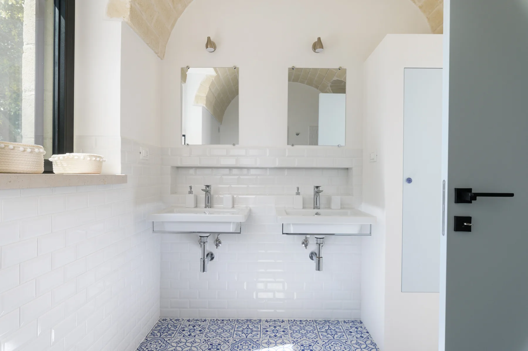 A bright bathroom features two sinks beneath mirrors and white tiled walls.