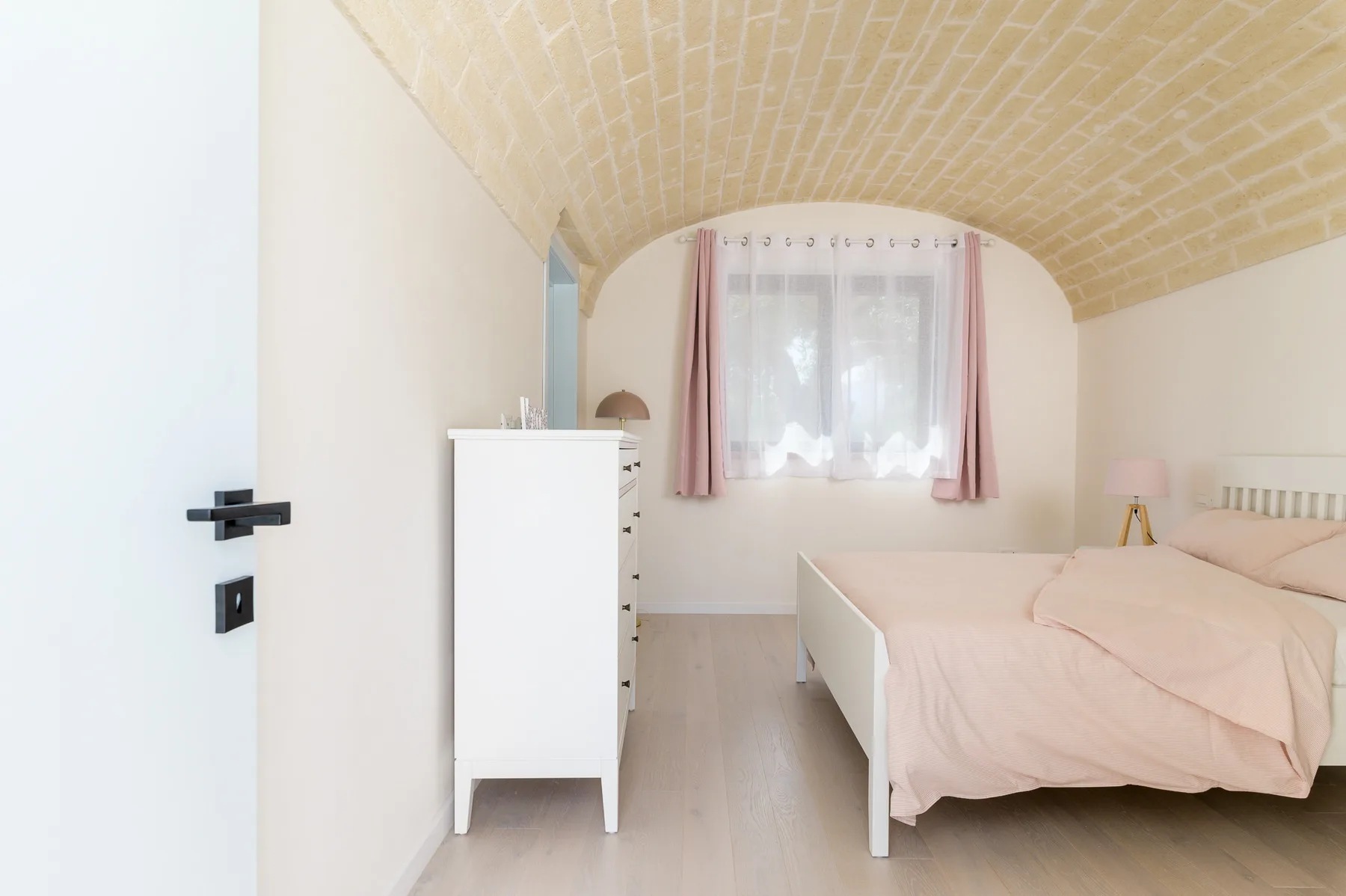 A bedroom with a bed beneath a curved brick ceiling and a curtained window.