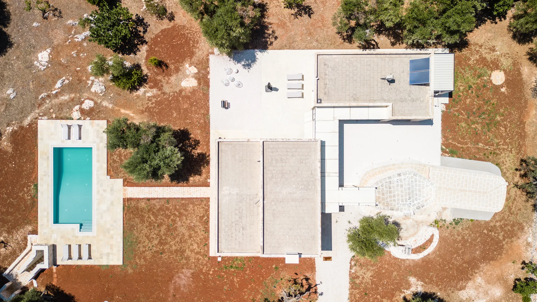 An aerial view of a modern building with a swimming pool set in dry, tree-dotted grounds.