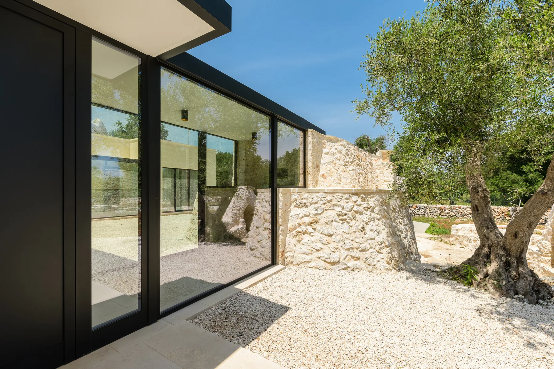 A modern glass-fronted building opens onto a bright gravel courtyard with trees.