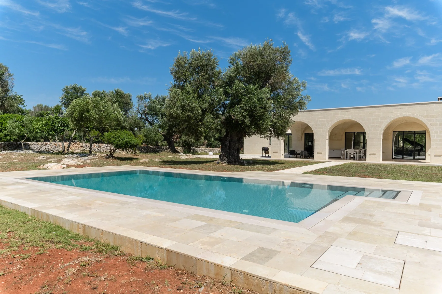 An outdoor swimming pool sits beside a low building with arched openings.