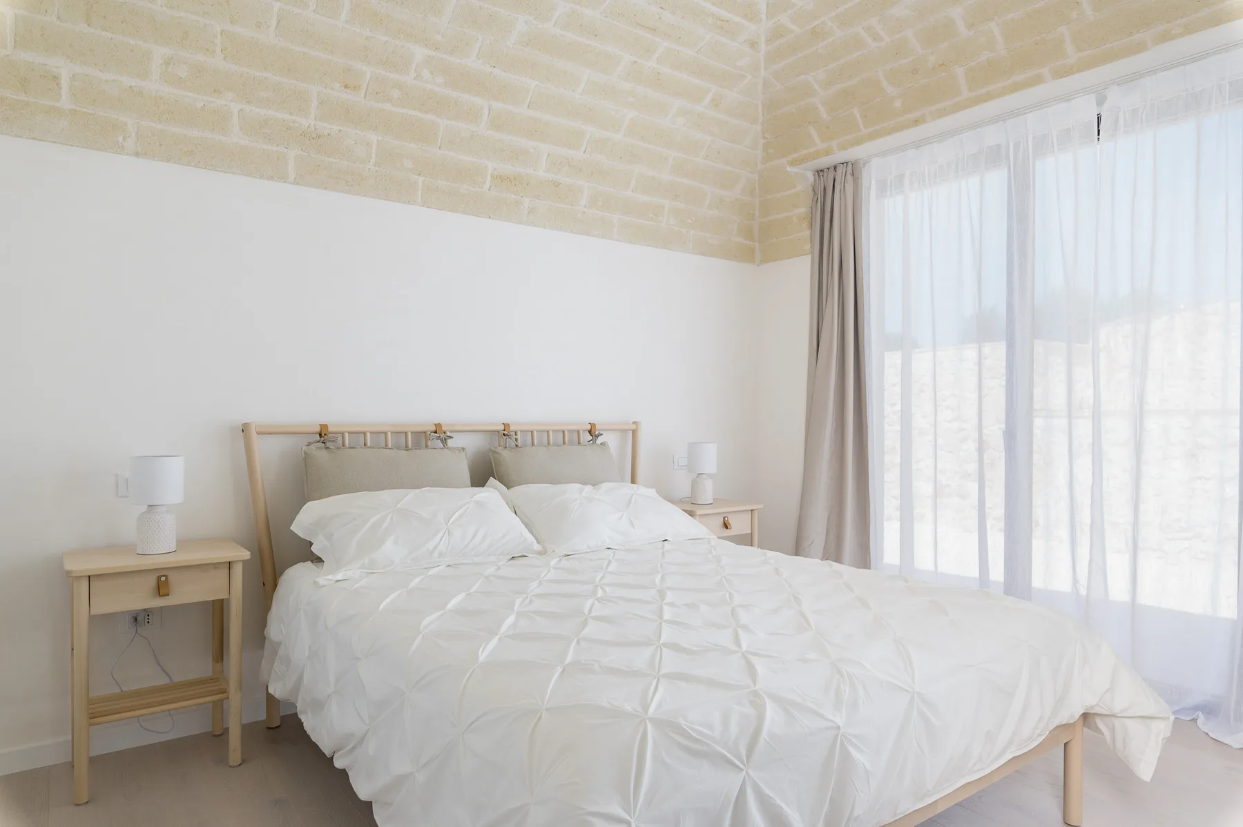 A bright bedroom with a bed dressed in white linens beside a large curtained window.