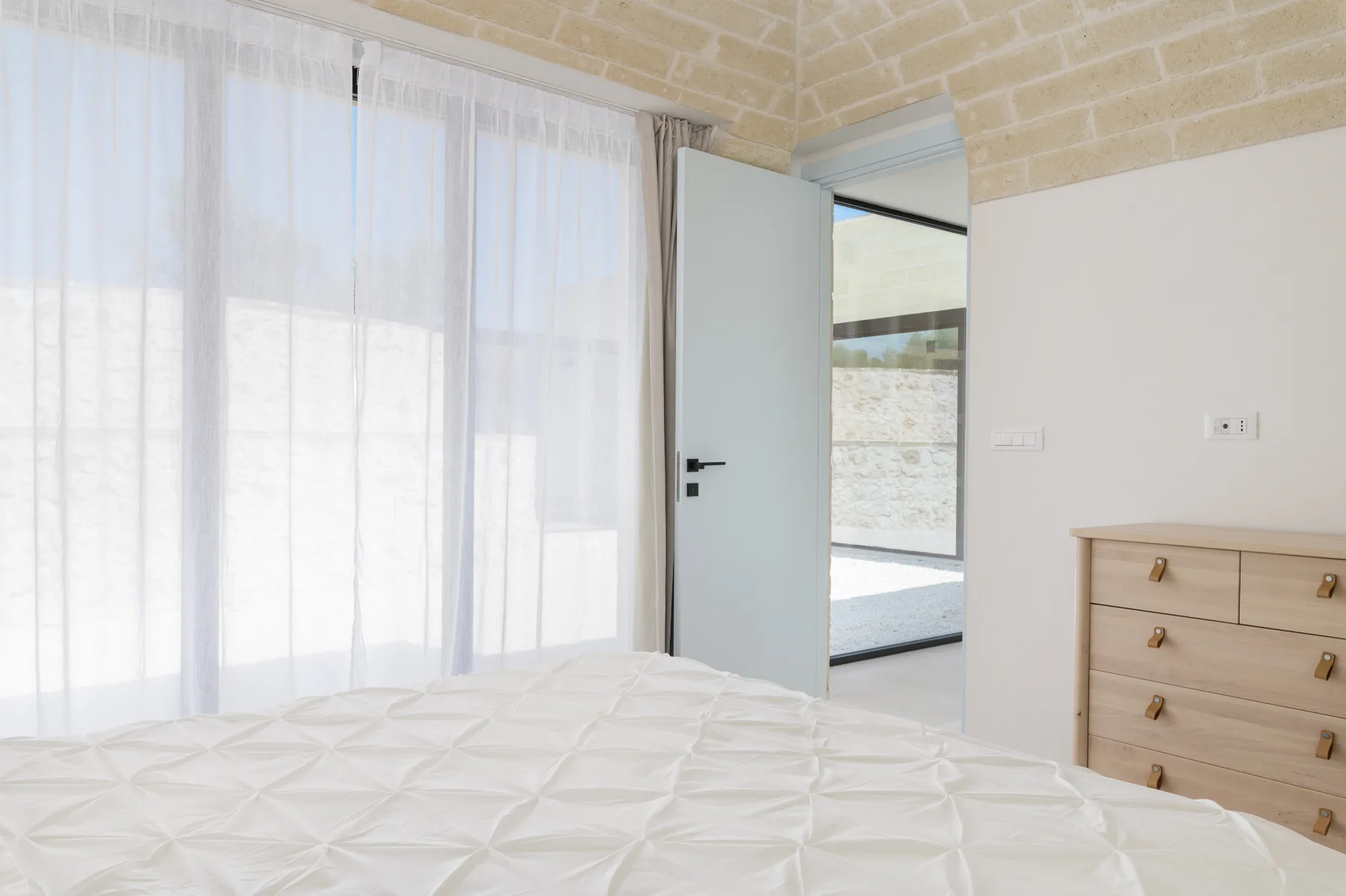 A bright bedroom with a bed beside an open door and sheer curtains.