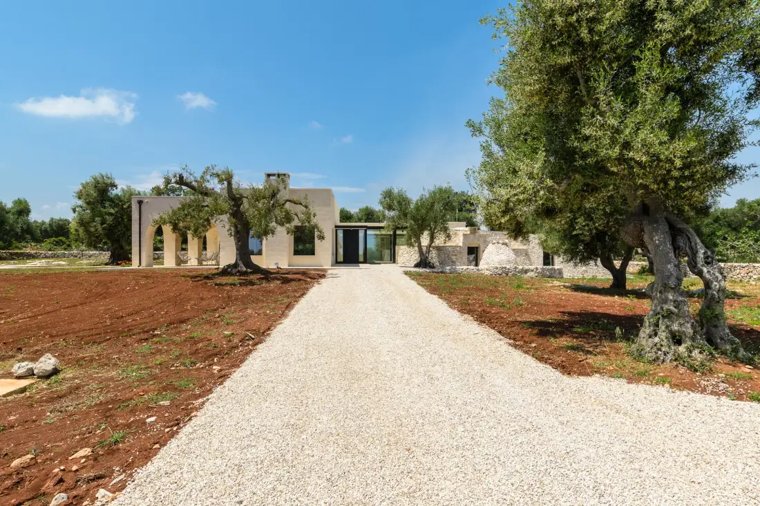 A modern building stands at the end of a pale gravel driveway, with trees on either side.