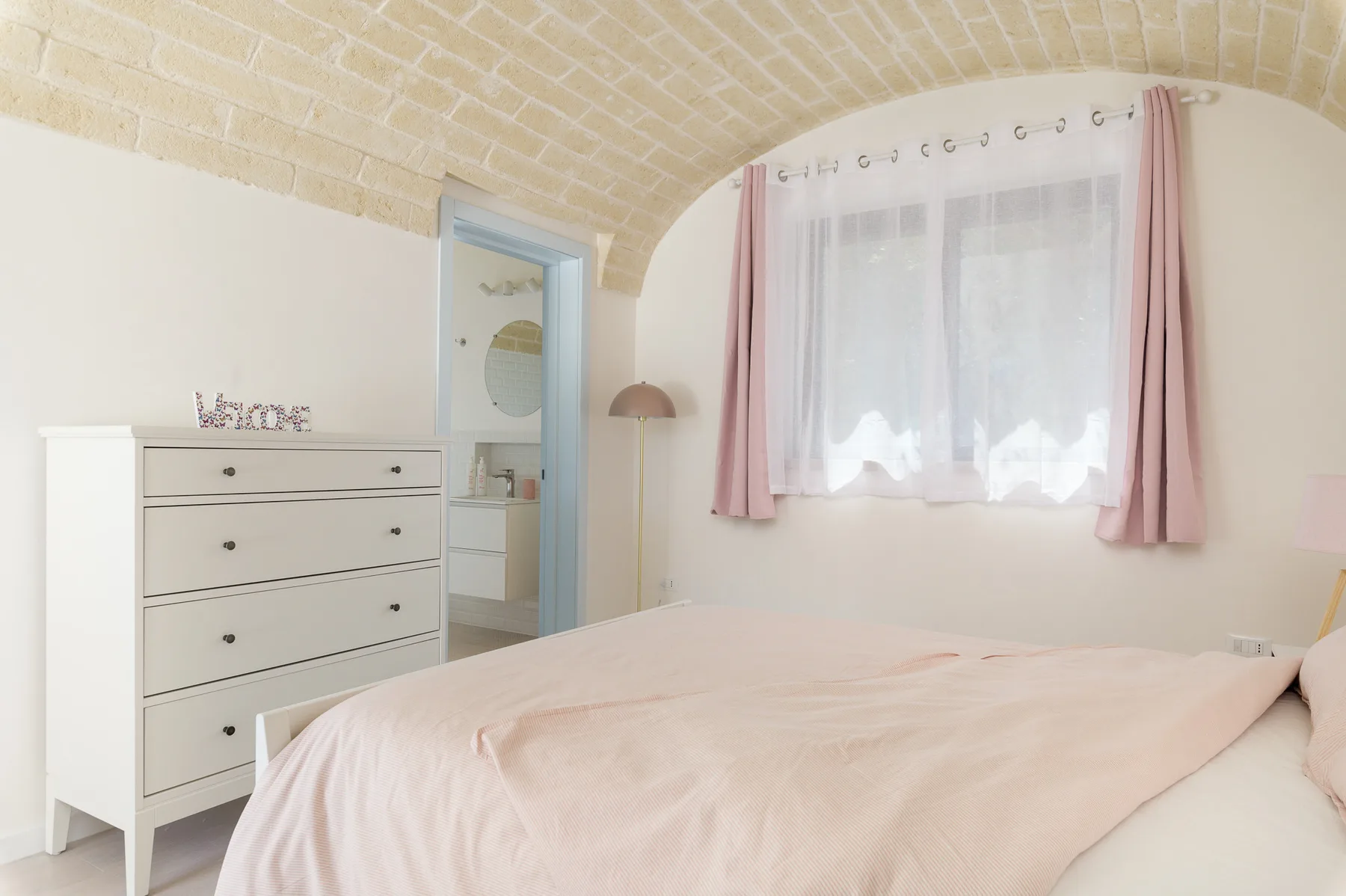 A bedroom with a pale bed, a white dresser, and a curtained window beneath a brick arch.
