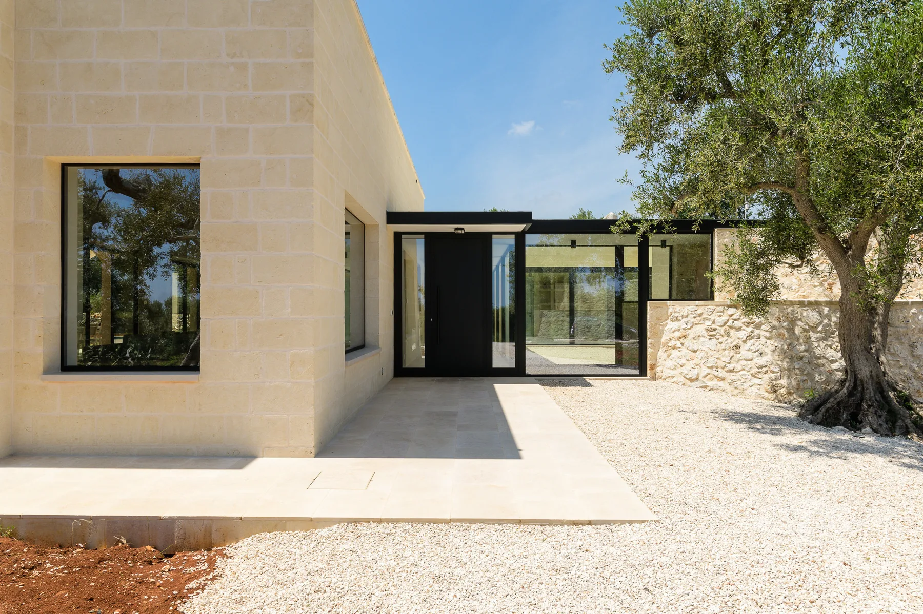 Modern house exterior with a glass entrance beside a gravel courtyard and tree.